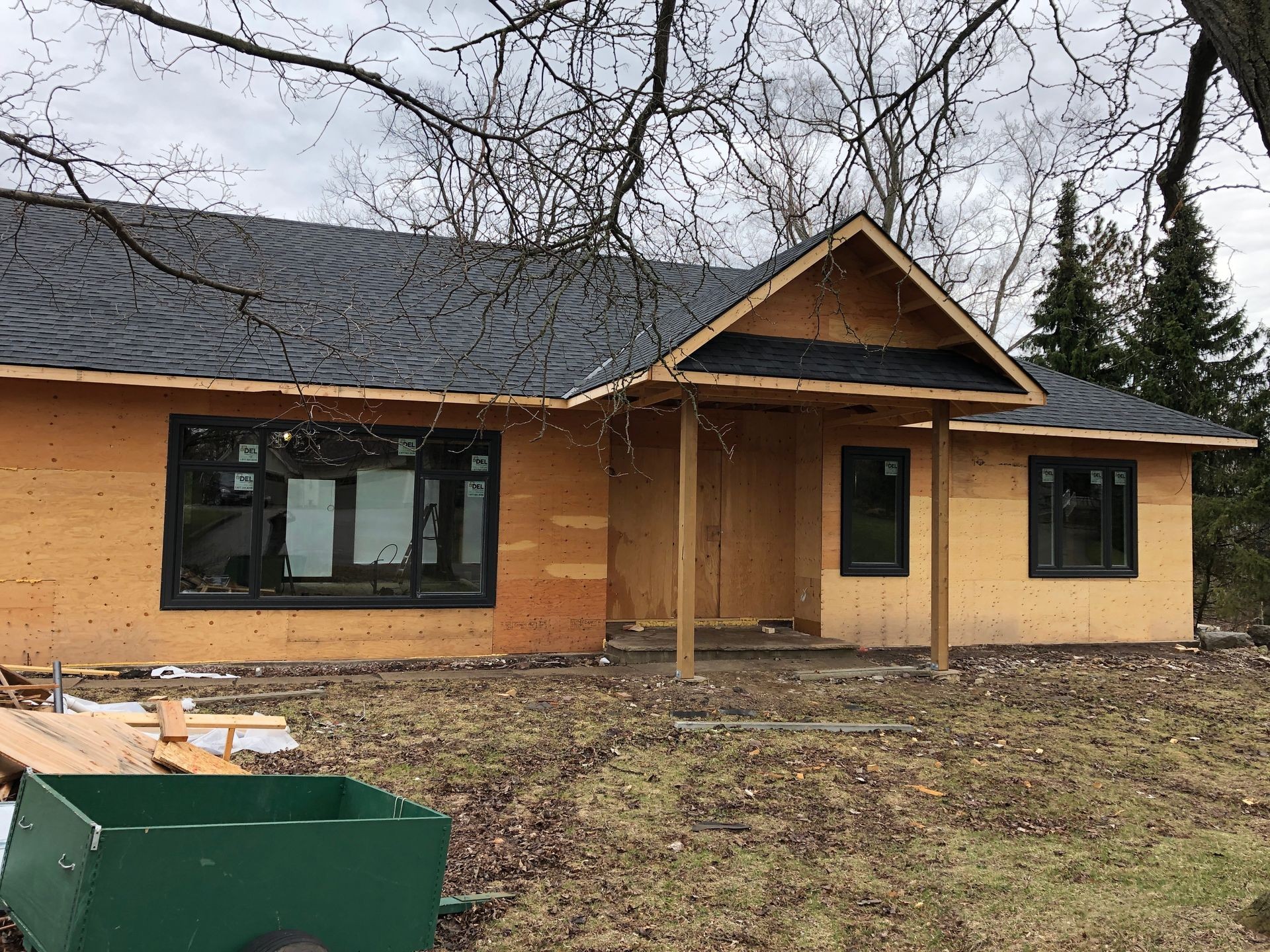 House under construction with plywood exterior sheathing and black framed windows. The structure features a pitched roof with dark shingles and a covered front porch supported by wooden posts. Construction materials and debris are visible in the foreground on a bare yard with sparse grass and leafless trees.