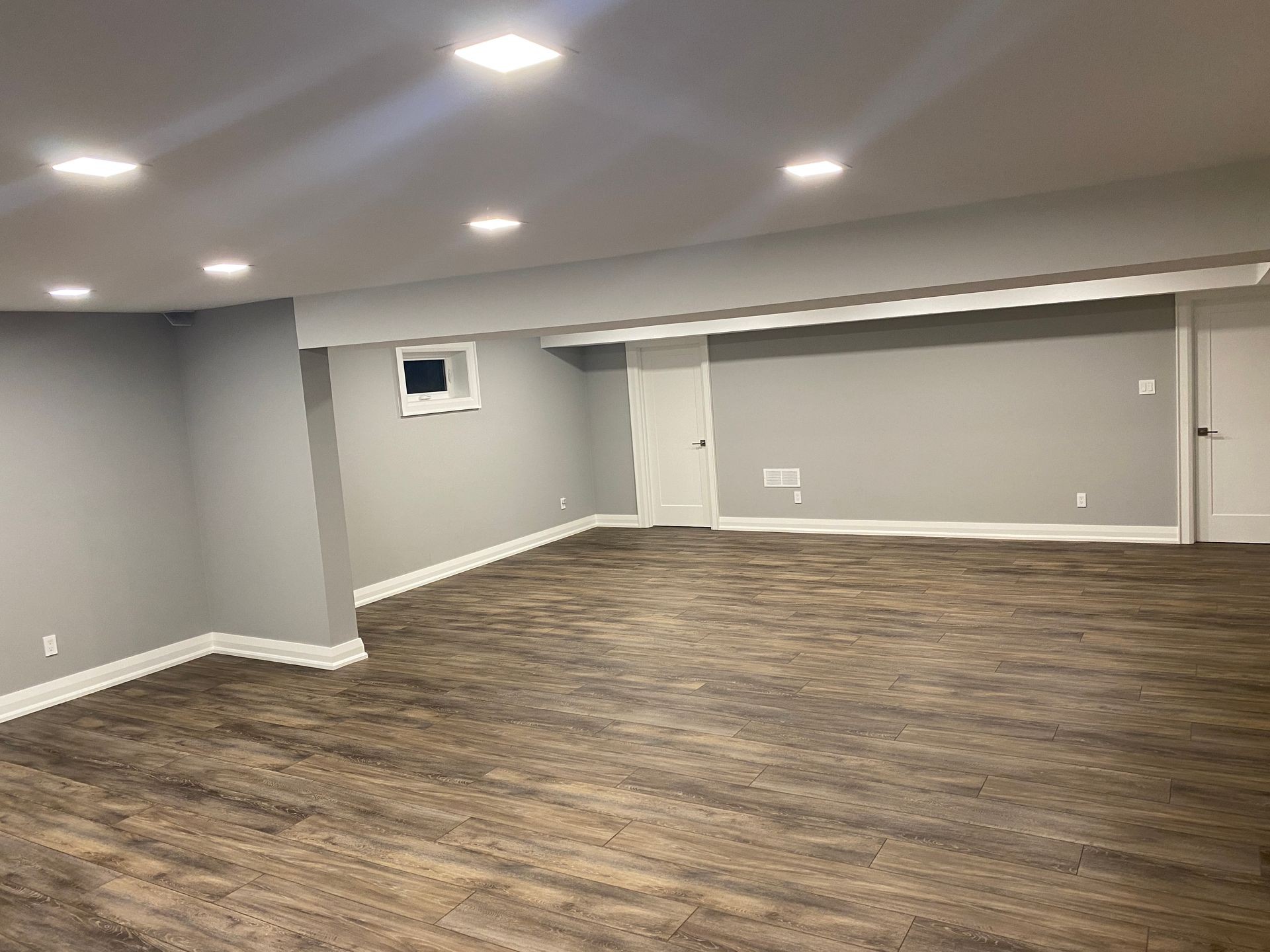 Modern finished basement with recessed ceiling lights, gray walls, and wide plank wood-look flooring. The space features clean white trim, two white doors, and a small window, creating a bright, open, and contemporary atmosphere.