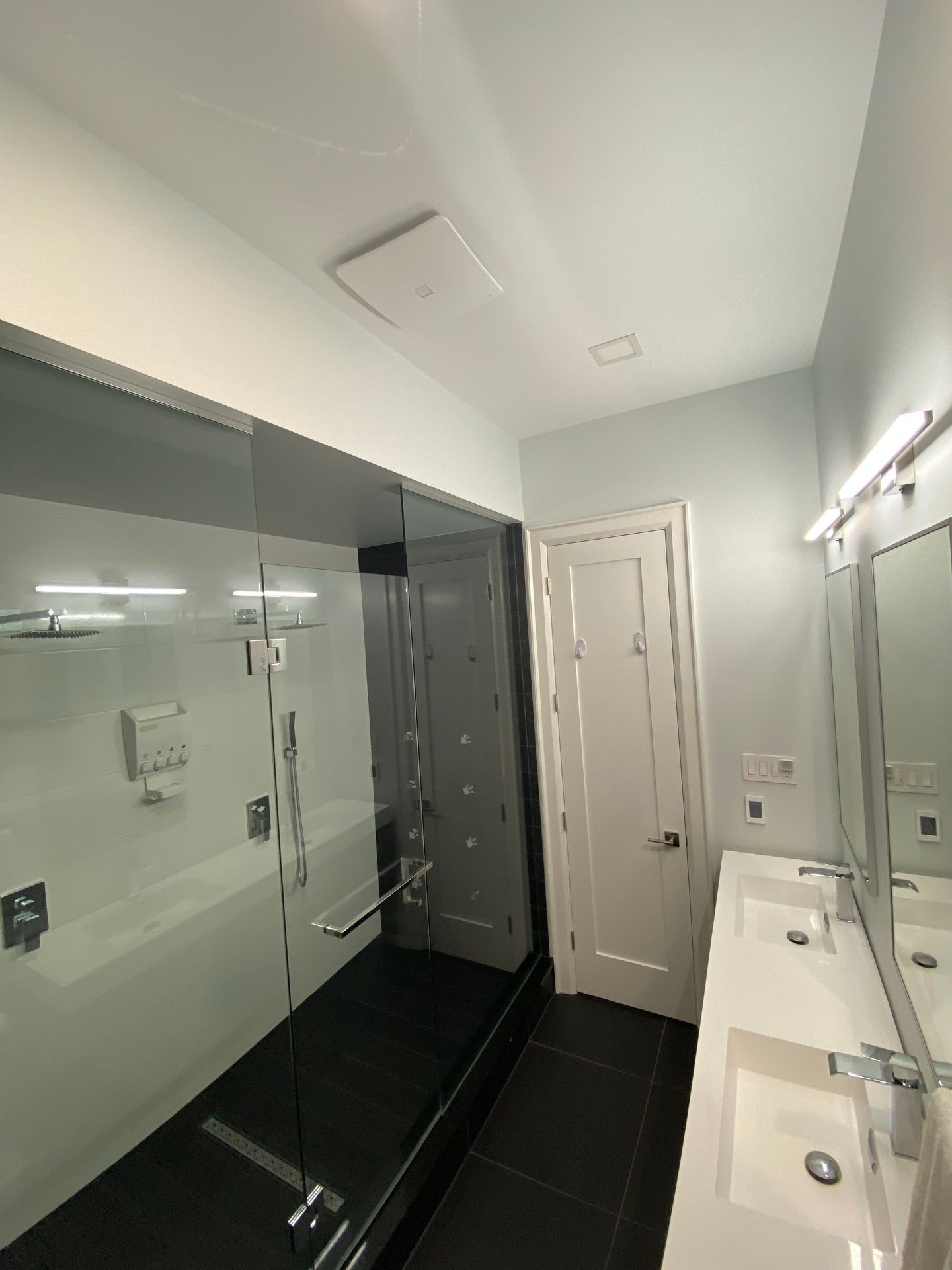 Contemporary bathroom featuring a glass-enclosed walk-in shower with black tile flooring and chrome fixtures. The vanity includes dual rectangular sinks with modern faucets, large mirrors, and sleek wall-mounted lights, creating a clean and minimalist design.