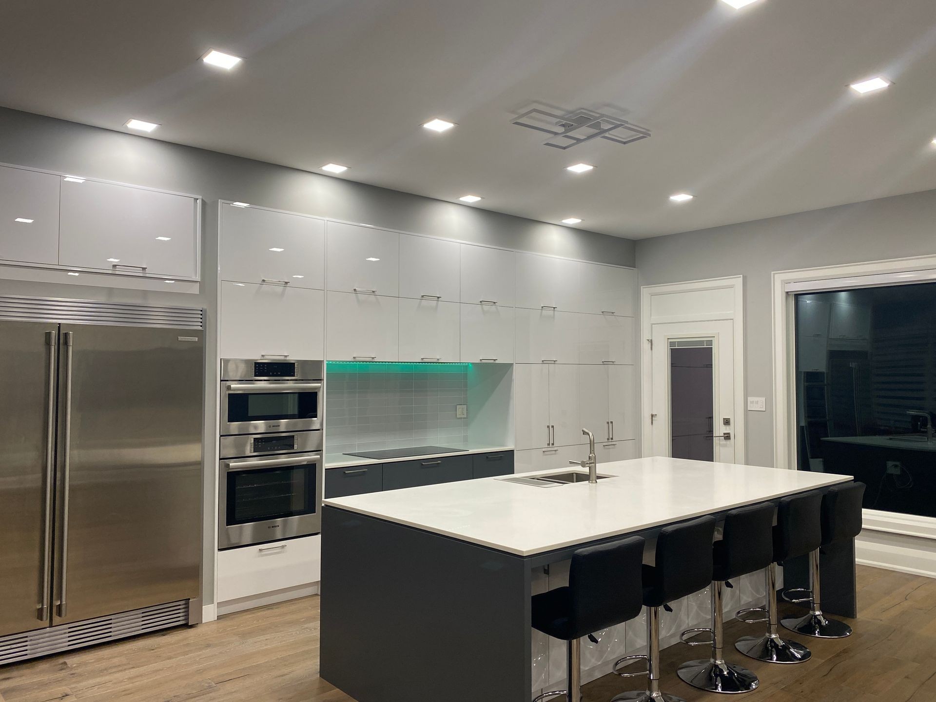 Modern kitchen with sleek white cabinets, stainless steel appliances, and a large island with seating for five. Features include a built-in double oven, integrated refrigerator, minimalist fixtures, under-cabinet lighting with a green accent, and recessed ceiling lights for a bright, contemporary look.
